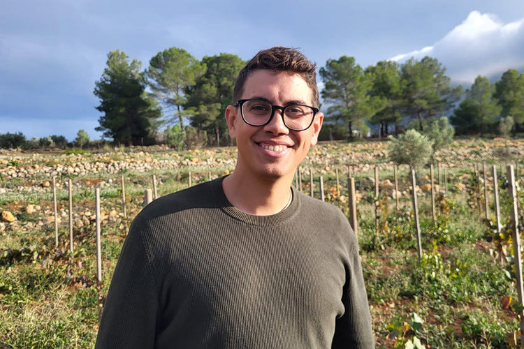 Persona sonriente con gafas de pasta y jersey verde posando en un viñedo, con fondo de parras, árboles y cielo parcialmente nublado.
