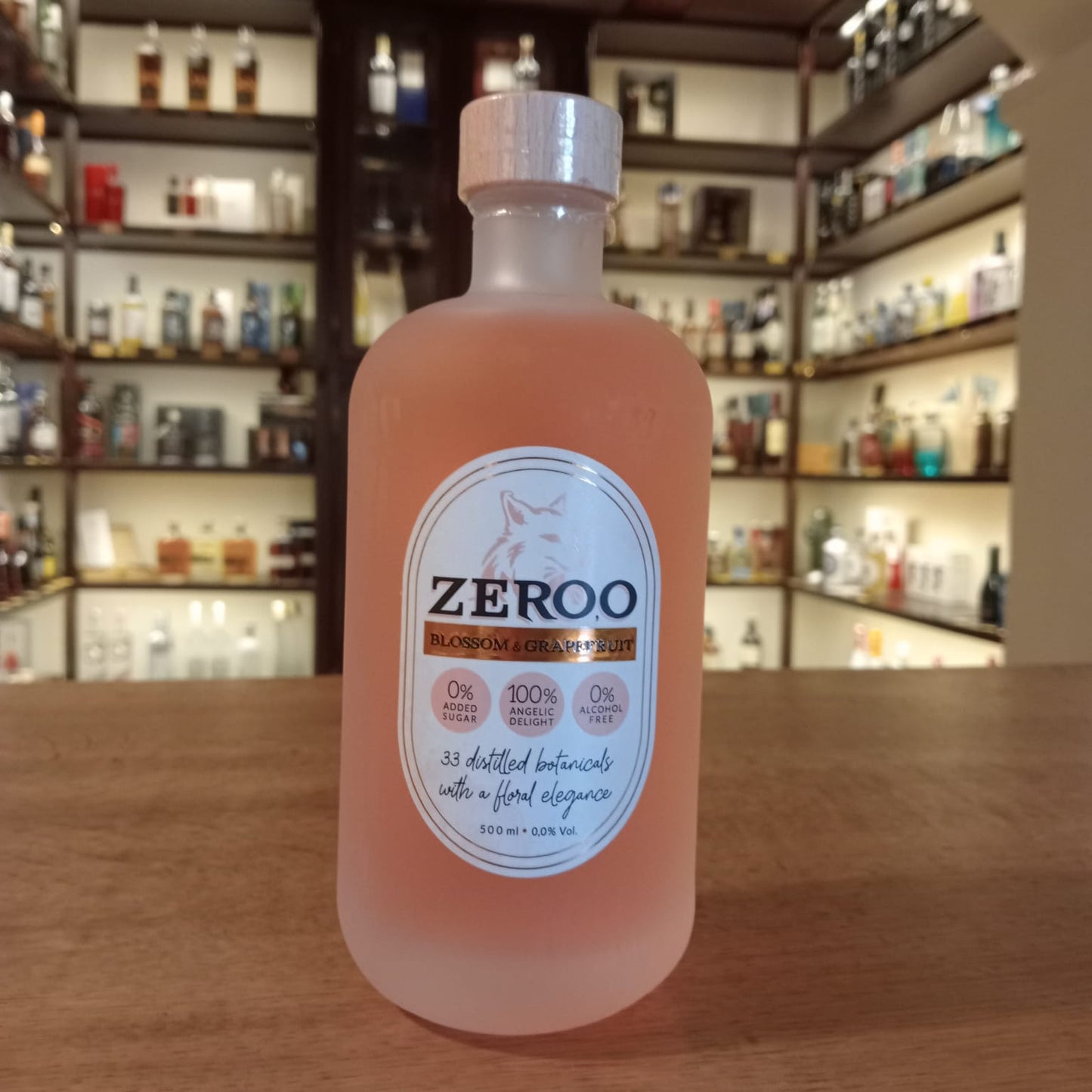 Botella de bebida Zeroo Blossom & Grapefruit sin alcohol, de color rosa suave en vidrio esmerilado, con etiqueta blanca y tapón de madera, colocada sobre mostrador de madera con estanterías de botellas al fondo.