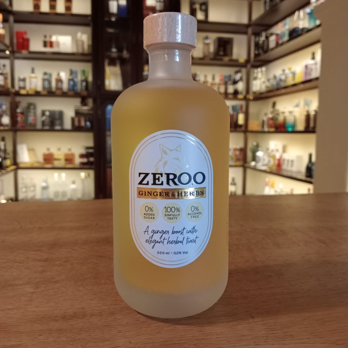 Botella de bebida Zeroo Ginger & Herbs sin alcohol, de color amarillo pálido y vidrio esmerilado, con etiqueta blanca y tapón de madera, colocada sobre mostrador de madera con estanterías de botellas al fondo.