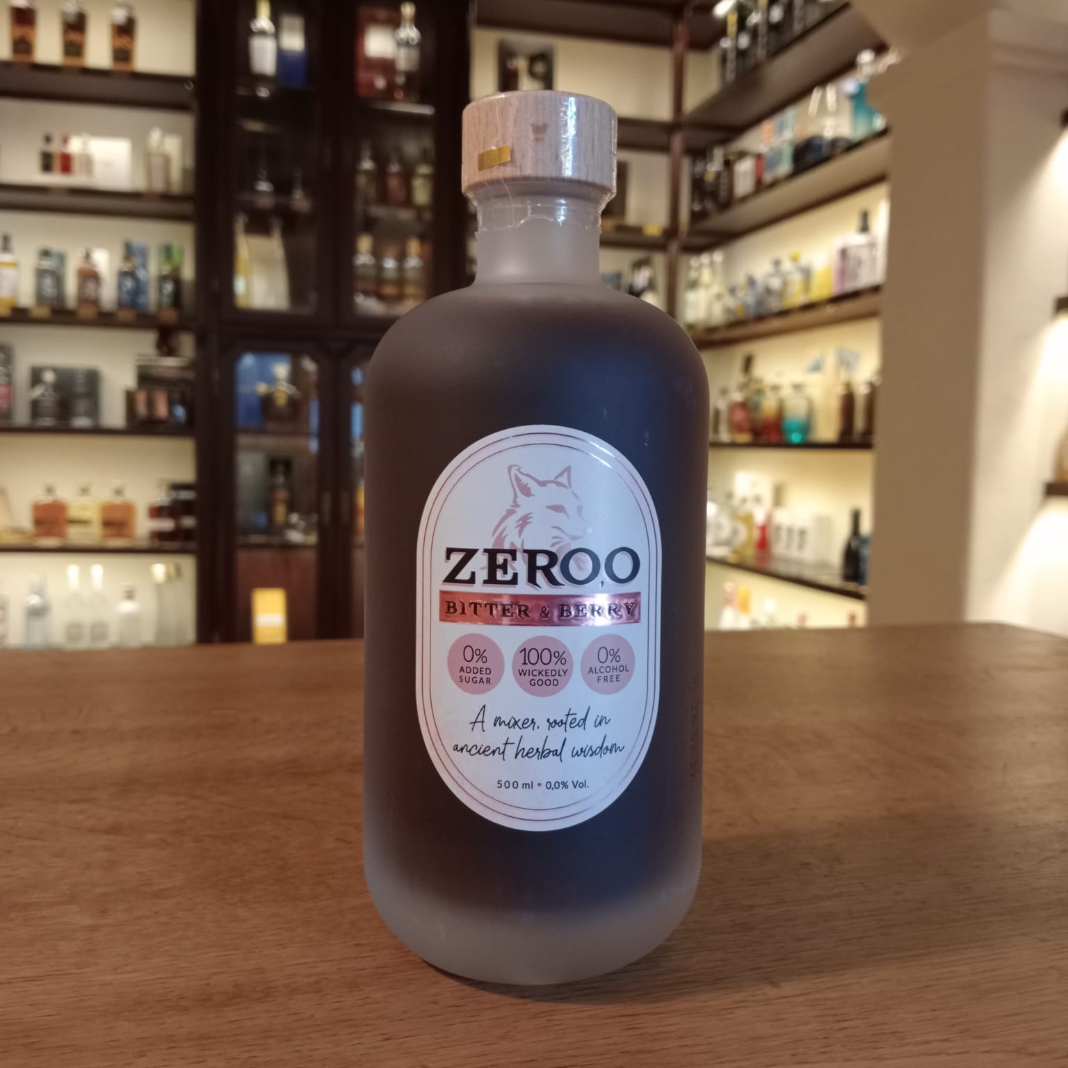 Botella de bebida Zeroo Bitter & Berry sin alcohol, de color rojo oscuro en vidrio esmerilado, con etiqueta blanca y tapón de madera, colocada sobre mostrador de madera con estanterías de botellas al fondo.