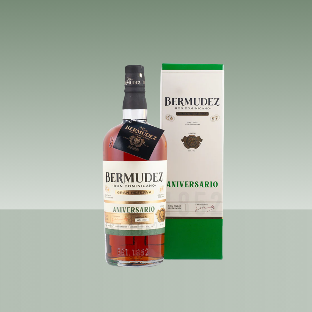 Botella de ron dominicano Bermúdez Aniversario Gran Reserva, con color ámbar, etiqueta clásica en tonos crema y verde, presentada junto a su estuche original sobre fondo neutro.