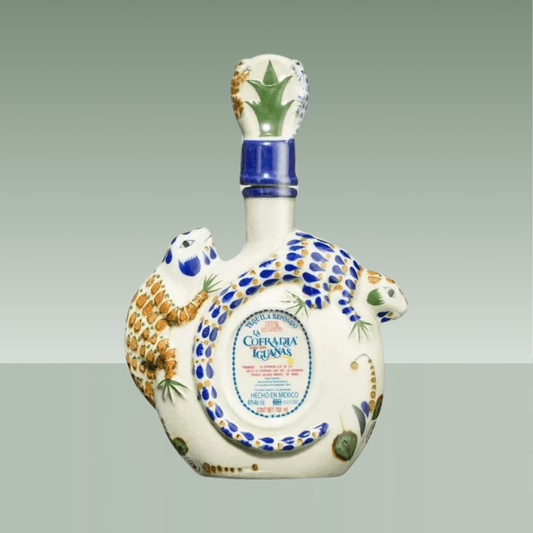Botella de tequila reposado de cerámica artesanal con forma de iguana, decorada a mano en blanco, azul y tonos ocres, con tapón cerámico y etiqueta frontal.