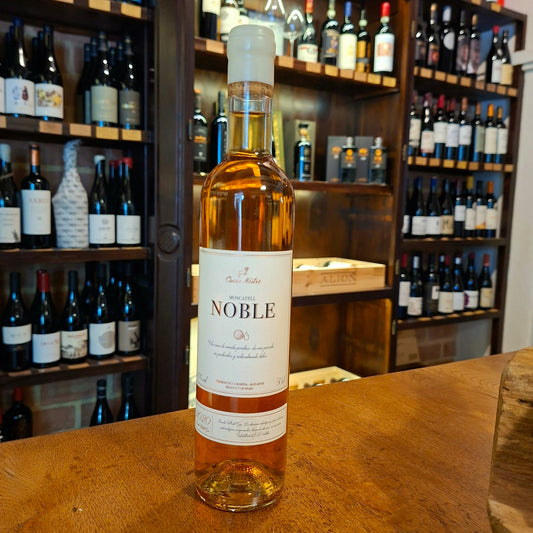 Botella de vino dulce Moscatel Noble de color ámbar, con etiqueta clara y cápsula blanca, expuesta sobre mesa de madera en interior de tienda.